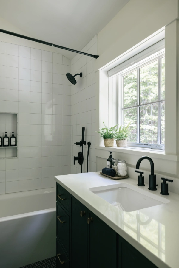 Bathroom vanity with matte black fixtures, white countertop, tiled shower surround, and window-side greenery